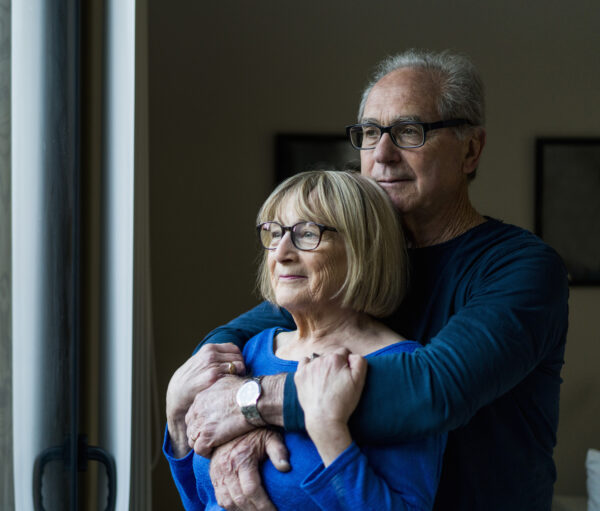 A photo of thoughtful senior couple looking through window. Loving man embracing woman at home. They are wearing casuals.